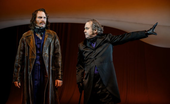 Två män ca 40–50 år i mörka rockar står mot röd bakgrund; den ene pekar åt sidan med handske.