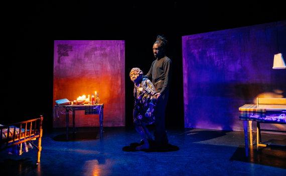 Man i svarta kläder står bakom en docklik äldre kvinna i blommig klänning på scen med säng och bord.