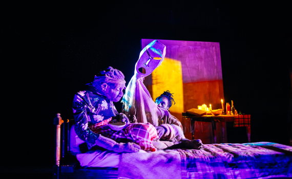 Docklik äldre kvinna sitter på säng medan en vuxen man bredvid håller upp en vit mask på en teaterscen.