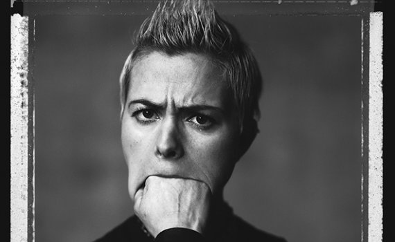 Kvinna med kort ljust hår och ärmlös svart tröja biter i handen, armtatuering synlig.