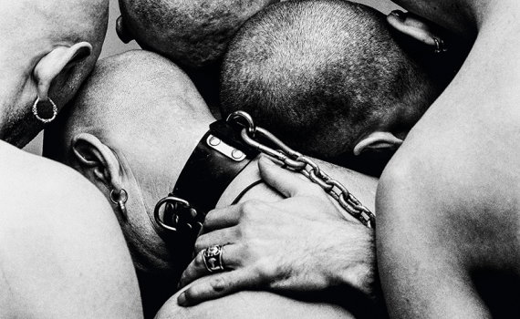 Fyra rakade personer tätt ihop, en med läderhalsband och kedja, närbild bakifrån i svartvitt.