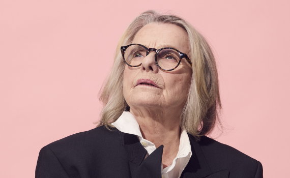 Kvinna i svart kavaj och glasögon ser uppåt mot något utanför bild. Porträtt mot en enfärgad rosa bakgrund.