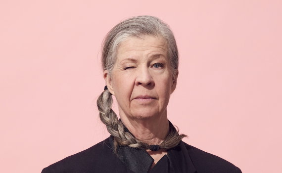 Person i svart kavaj med grå fläta sitter rakt fram och blinkar. Händerna är knäppta framför kroppen mot rosa bakgrund.