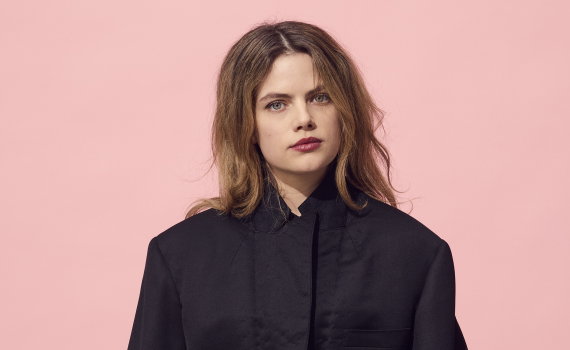 Ung kvinna i svart kostym sitter på en låda och ser rakt mot kameran. Studioporträtt mot rosa bakgrund.