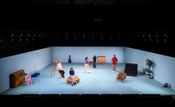Helbild av scenrum med fem personer utspridda bland stege, piano, fåtölj och andra objekt.