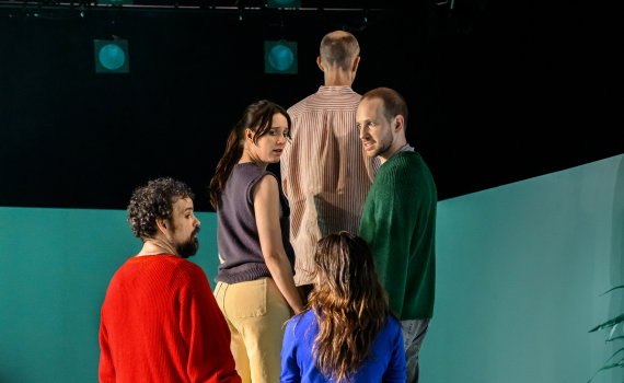 Fem personer står tätt tillsammans på en upphöjd plattform och ser ut mot publiken under scenljus.