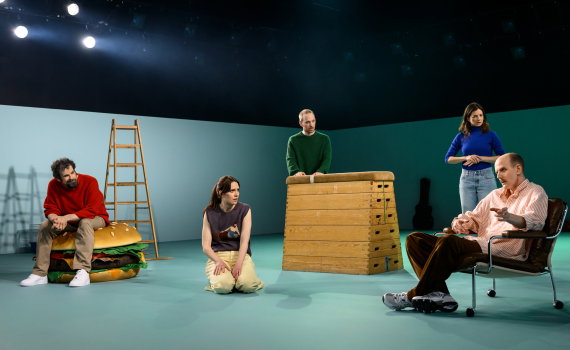 Fem personer i samtal på en öppen scen; en sitter i stol och gestikulerar, andra står eller sitter kring rekvisita som stege och låda.