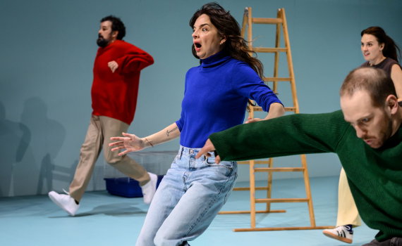 Tre personer i rörelse på scen; kvinna i blå tröja i fokus, andra springer förbi i bakgrunden.