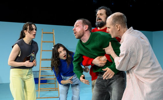 Fyra personer håller i en man i grön tröja som lutar framåt; uttrycksfull scen med dramatisk energi.