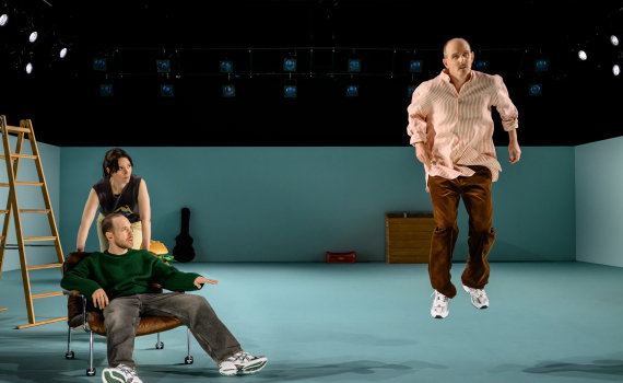Man i randig skjorta svävar i luften medan två personer ser på; scen med stege och rekvisita.