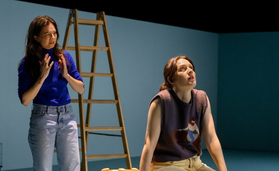 Två kvinnor vid stor hamburgarrekvisita; en sitter och ser upp, den andra står bakom med händerna lyfta.