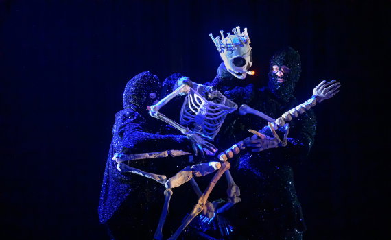 Två vuxna personer i svarta, glittriga helkroppsdräkter håller en skelettdocka med krona på mörk scen.