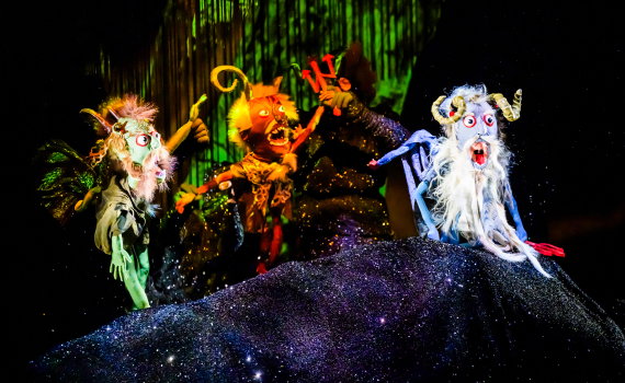 Tre färgglada djurdockor med horn och vingar framförs av svartklädda vuxna på upplyst scen.