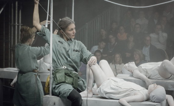 Kvinna ca 25–35 år i grön arbetsdräkt håller i trådar fästa vid ett barn målat vitt som ligger på ett bord, publik i bakgrunden.