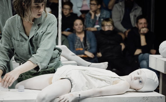 Kvinna ca 25–35 år i grön arbetsdräkt arbetar vid ett bord där ett barn målat vitt ligger på rygg, publik sitter bakom.