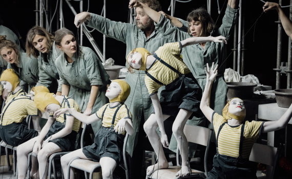 Fyra vuxna i gröna arbetsdräkter styr barn målade vita med trådar som marionetter på stolar, scenutrustning bakom.