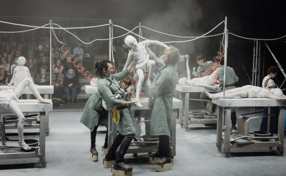 Scenföreställning där tre vuxna personer i gröna arbetskläder lyfter ett barn målat vitt på ett bord, publik sitter bakom.