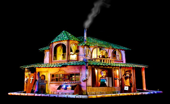 Färgglad modell av ett hus med flera rum där docklika figurer syns i fönster och på balkong, rök stiger från skorstenen.
