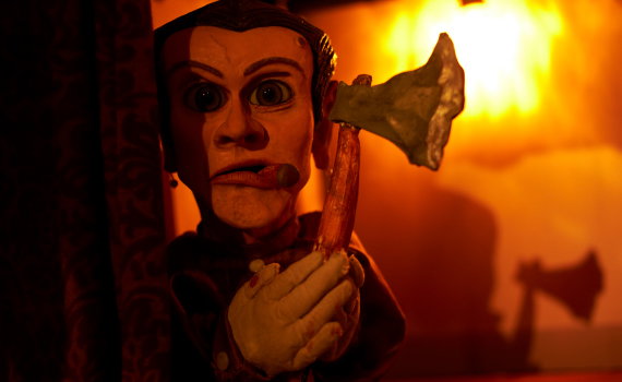 Dockfigur i röd belysning håller en yxa, dramatisk scen med starka skuggor på väggen bakom.