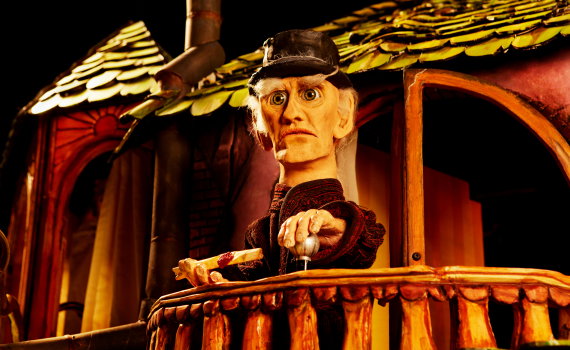 Dockfigur med hatt står på en balkong och håller i en spak, upplyst i varmt ljus mot husfasaden.