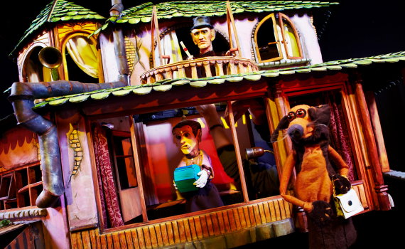 Interiör av modellhus med flera dockfigurer; en håller en låda medan en hundlik figur står i förgrunden.