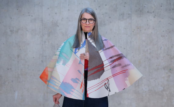 Kvinna med glasögon bär färgstark cape och håller tygstycke, står mot neutral betongvägg.