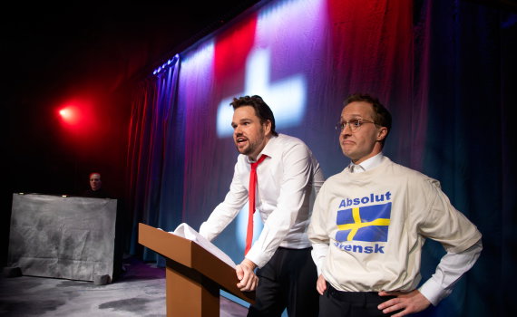 Helan som amerikansk pastor vid ett podie och Halvdan som översättare bredvid honom.