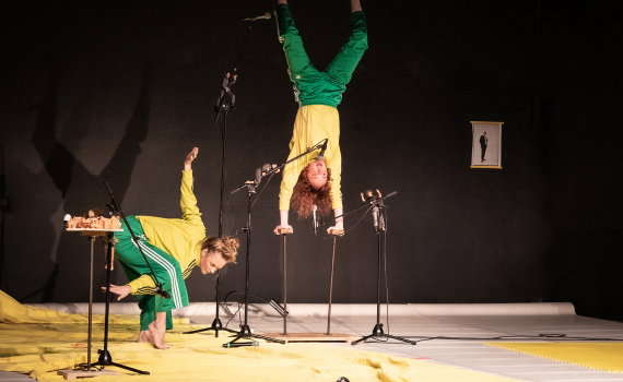 Två performers i gröna och gula träningskläder uppträder på scen med mikrofoner och akrobatiska rörelser.