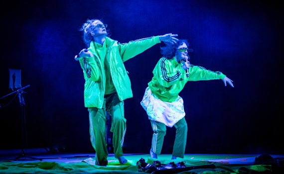 Två performers i gröna scenkläder sjunger och gestikulerar på en blåbelyst scen med mikrofon i handen.