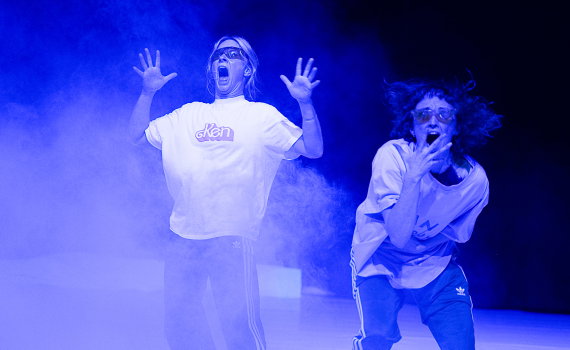 Två performers i vita t-shirts och solglasögon reagerar dramatiskt i blått scenljus och rök.
