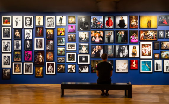 Besökare sitter på en bänk framför en stor vägg fylld med fotografiska porträtt.