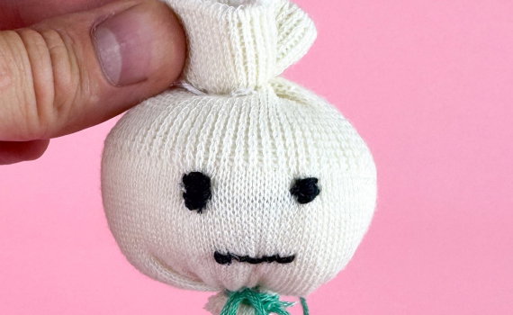 En hand håller upp en sydd vit lökfigur med broderat ansikte mot en rosa bakgrund.