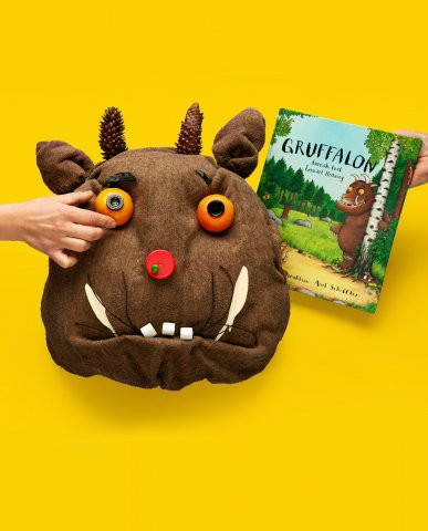 Ett monsterliknande huvud med orange ögon, röd näsa och betar, i bakgrunden boken Gruffalon. 