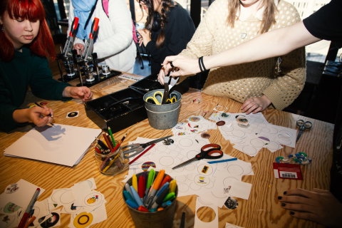 Grupp vid ett bord skapar klistermärken, med saxar, pennor och papper utspridda.