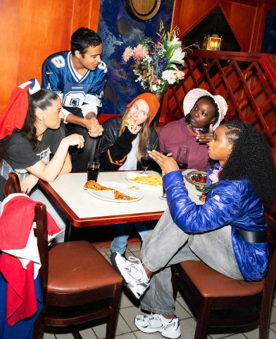 Fem unga personer sitter vid ett bord med mat, pratar och äter i en restaurangmiljö.