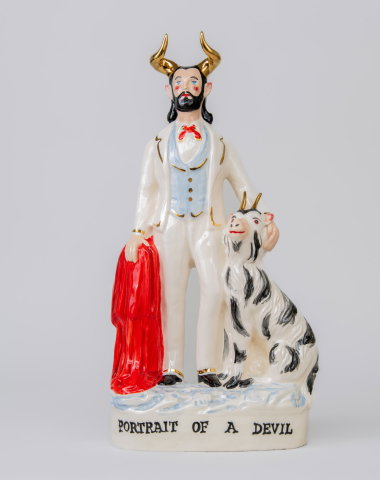 Porslinsfigur av en man med horn, skägg och kostym, hållande en röd mantel bredvid en getliknande varelse med horn.