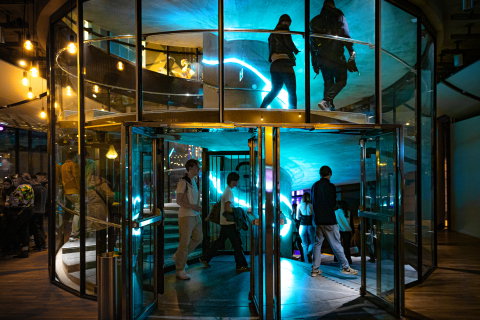 Flera personer går in genom glasad karuselldörr till en byggnad med neonljus.