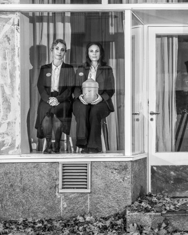 Helen Sjöholm och Anna Fahlstedt sitter på varsin pall i ett tomt skyltfönster. I sina händer har Anna en begravningsurna.