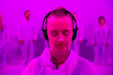 Kvinna i 30-årsåldern med ljust uppsatt hår och hörlurar, vit hoodie, blundar i rosa ljus, suddiga personer i vitt bakom.