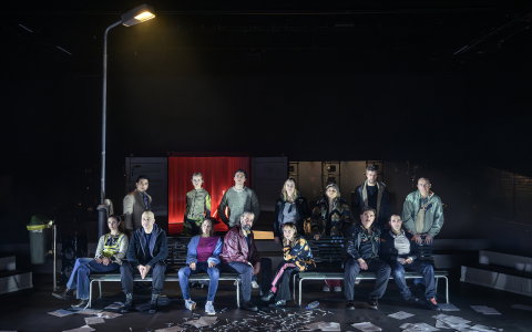 Tretton vuxna män och kvinnor i olika åldrar sitter och står på bänkar under gatlykta; sprutor och papper på golvet.