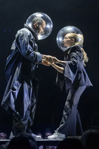 Man och kvinna i mörka kläder och stora genomskinliga hjälmar håller varandras händer på scen.
