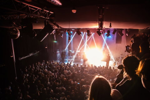 Band med flera musiker och trummor på scen i starka strålkastare; stor publik av vuxna står tätt framför scenen i konsertlokal.