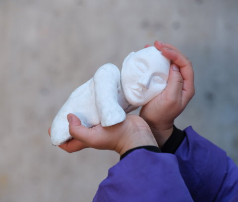 Ett person håller en liten vit skulptur av en liggande figur med slutna ögon i sina händer. Närbild mot en mjuk, neutral bakgrund.