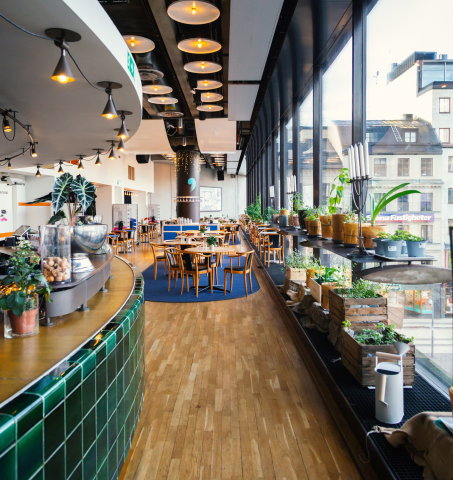 Ljus restaurang med trägolv, runda bord och stolar. Stora fönster längs väggen med krukväxter och utsikt över stadsgata.