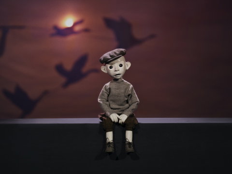 Docklik figur med basker sitter på en kant i dämpat ljus, med skuggor av flygande figurer projicerade på väggen bakom.