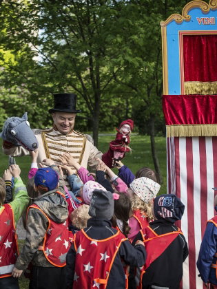 En dockskådespelare håller i en docka som ser ut som en varg och en docka som ser ut som rödluvan, en grupp barn tittar på. 