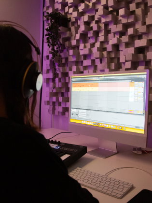 Person med hörlurar vid dator i musikstudio med lila ljus och ljudpanel på väggen.