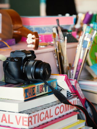 Närbild av kamera, penslar, garn och staplade böcker på ett bord.
