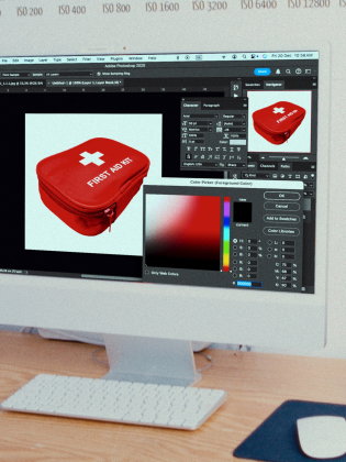 En datorskärm som visar ett designprogram med en röd förstahjälpen-väska. På bordet finns kamera, tangentbord och mus.