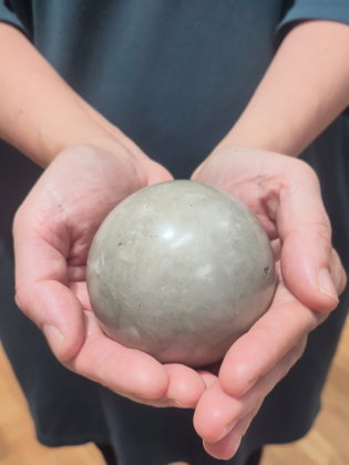 Händer som håller en rund, grå stenboll med slät yta, personen bär en mörkblå tröja i bakgrunden.